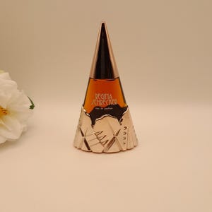 Puede incluir: Un frasco de perfume con un diseño único. El frasco tiene forma cónica con una parte superior dorada y una base plateada. Contiene un líquido marrón y tiene el texto "REGINA SCHRECKER eau de parfum".