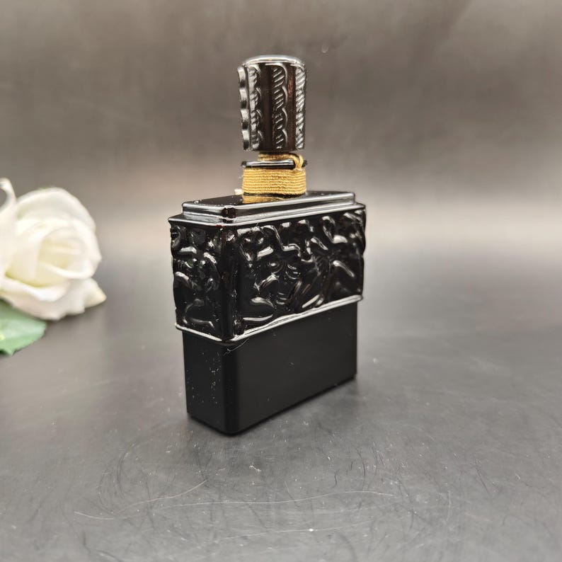 May include: A black perfume bottle with a decorative stopper. The bottle has a rectangular base and a detailed relief design. The stopper is silver and black, and the neck is wrapped with gold-colored twine. A white rose is in the background.