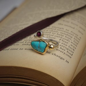 May include: A ring featuring a polished turquoise stone set in a gold-toned bezel, accented by a small round red gemstone and a smooth gold-toned sphere on a silver-toned band. The ring is positioned on the open pages of a printed book.