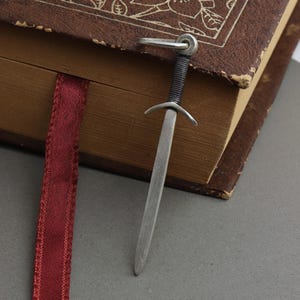 May include: A silver sword bookmark with a black wrapped handle is inserted into a brown book. A burgundy ribbon bookmark is visible. The bookmark is made of metal and has a detailed design. The book has a worn appearance.
