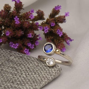 May include: A silver bypass ring featuring a round blue gemstone in a bezel setting, a smaller round clear gemstone in a bezel setting, and a small gold bead accent. The ring is positioned near a cluster of small purple flowers.