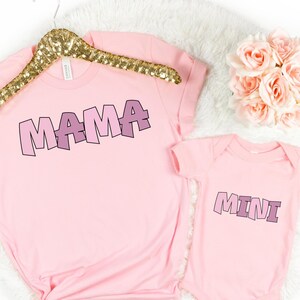 May include: Pink t-shirts with the words "MAMA" and "MINI" in a playful, layered font. The shirts are perfect for a mother and child.