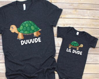May include: Two dark gray t-shirts with turtle graphics. One shirt says "DUUUDE" and the other says "LIL DUDE". The turtles are green and brown. The shirts are on a light brown wooden surface.