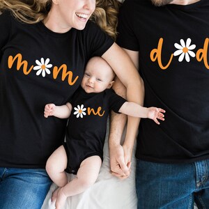 May include: A family of three wearing matching black t-shirts with orange text and white daisy designs. The shirts say "mom", "dad", and "one".