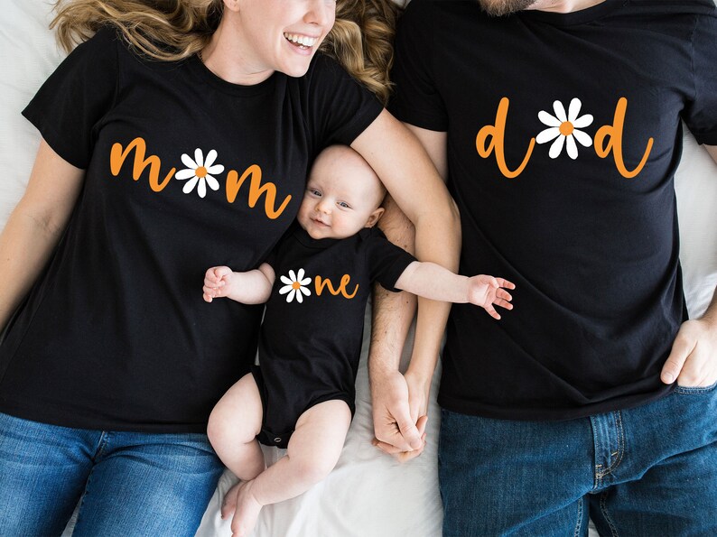 May include: A family of three wearing matching black t-shirts with orange text and white daisy designs. The shirts say "mom", "dad", and "one".