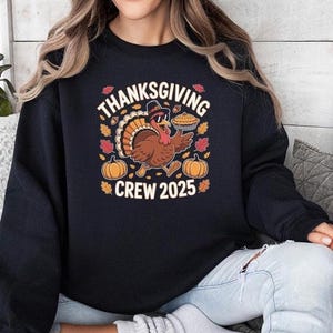 May include: Black sweatshirt with a Thanksgiving-themed graphic. The design features a turkey wearing sunglasses and a hat, holding a pie, surrounded by pumpkins and fall leaves. Text reads "THANKSGIVING CREW 2025".