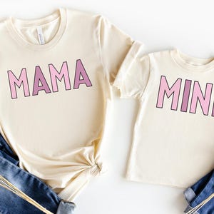 May include: Two cream colored t-shirts with pink and purple ombre lettering. The larger shirt says "MAMA" and the smaller shirt says "MINI".