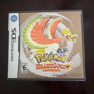 May include: A Nintendo DS game case for Pokémon HeartGold Version. The cover features a stylized orange and yellow bird with green accents. The game title is in a gold font. The case has the Nintendo DS logo and Wi-Fi connection icon.