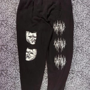 May include: Black sweatpants with a drawstring waist. The left leg features two white, cracked-face mask graphics. The right leg has three vertical white text graphics. The sweatpants are made of a soft material.