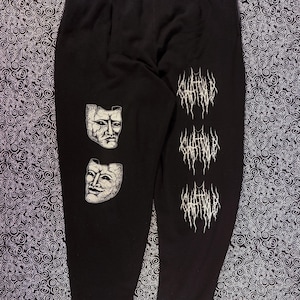 May include: Black sweatpants with a drawstring waist. The left leg features two white, cracked-face mask graphics. The right leg has three stacked white text graphics with a gothic font. The sweatpants are displayed on a black and white patterned surface.