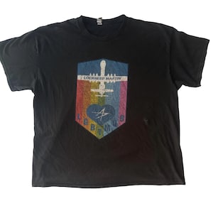Può includere: T-shirt nera con un design grafico colorato. Il design presenta una forma a scudo con un aereo, un cuore e il testo "LOCKHEED MARTIN" in alto. La parte inferiore dello scudo riporta le lettere "GBMO".