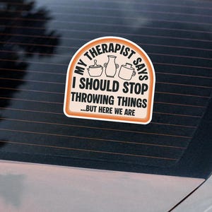 以下が含まれることがあります： 車の窓に貼られた白とオレンジ色のステッカー。「MY THERAPIST SAYS I SHOULD STOP THROWING THINGS...BUT HERE WE ARE」の文字と陶器のイラストが描かれています。