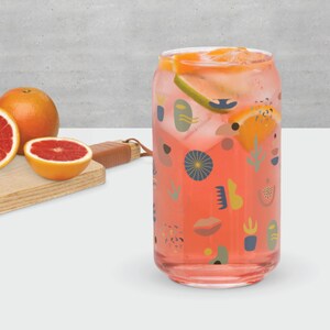 May include: A clear glass can-shaped drinking glass filled with a pink beverage, ice, and citrus slices. The glass is decorated with colorful abstract shapes. Grapefruit halves and whole grapefruit are on a wooden cutting board.