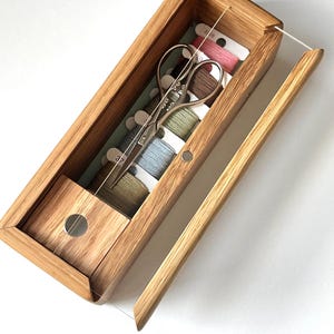 May include: Wooden thread organizer with a clear lid, containing spools of thread in various colors and a pair of small scissors. The wooden box is rectangular with a hinged lid and a small compartment. The thread colors include pink, brown, and blue.