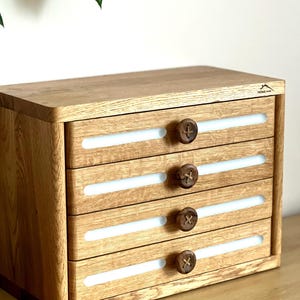 Könnte beinhalten: Holz-Aufbewahrungstruhe mit vier Schubladen. Jede Schublade hat einen weißen Akzent und einen runden Holzknopf. Die Truhe ist aus hellem Holz mit natürlicher Oberfläche gefertigt. Sie steht auf einer Holzoberfläche.