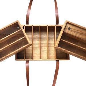 May include: Wooden organizer with multiple compartments and two hinged side compartments. The organizer has a natural wood finish and brown leather straps for carrying. The side compartments have shelves for storage. The organizer is designed for carrying and organizing items.