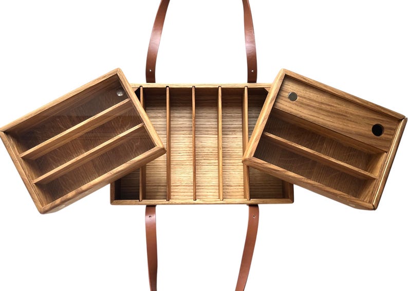 May include: Wooden organizer with multiple compartments and two hinged side compartments. The organizer has a natural wood finish and brown leather straps for carrying. The side compartments have shelves for storage. The organizer is designed for carrying and organizing items.