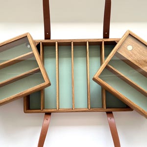 Pode incluir: Vitrine de madeira com compartimentos e prateleiras cobertas de vidro. O interior é verde claro e é suspenso por tiras de couro castanho. A vitrine abre em ambos os lados, revelando o interior.