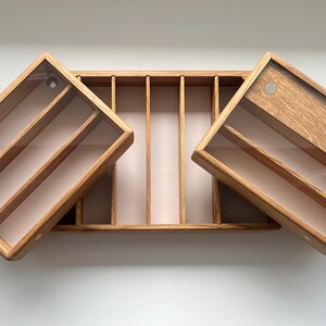 May include: Wooden organizer with a light brown finish. The organizer has a rectangular shape with a central compartment and two hinged, angled compartments. Each compartment has a clear lid and interior dividers. The organizer is on a white surface.