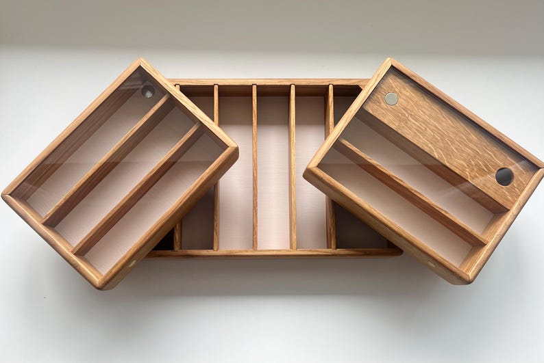 May include: Wooden organizer with a light brown finish. The organizer has a rectangular shape with a central compartment and two hinged, angled compartments. Each compartment has a clear lid and interior dividers. The organizer is on a white surface.