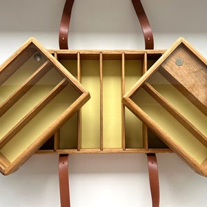 May include: Wooden storage box with a hinged lid and a brown leather strap. The box has a light wood finish with a yellow interior and clear glass panels. The box is open, revealing multiple compartments.