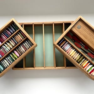May include: Wooden thread organizer with multiple compartments. The open box reveals colorful spools of thread and embroidery tools. The box is made of light-colored wood and has a light green interior.