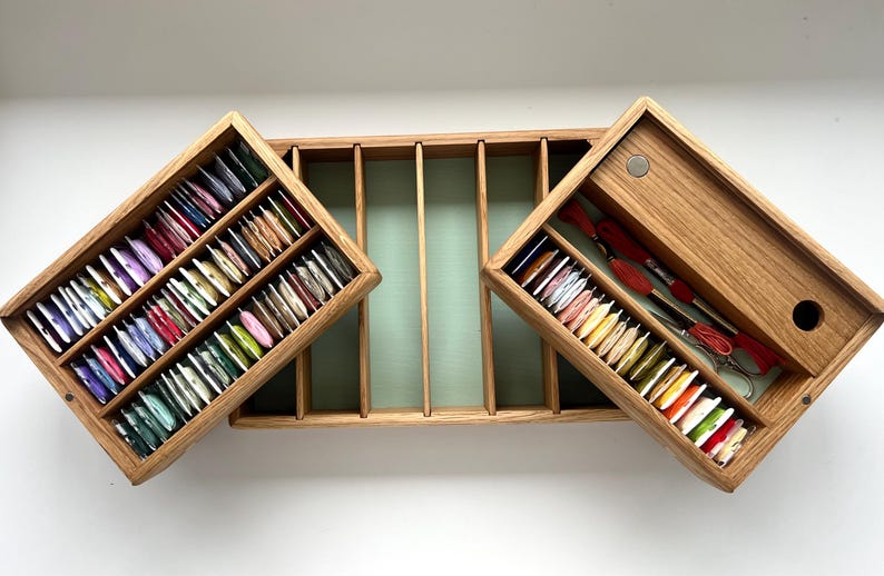 May include: Wooden thread organizer with multiple compartments. The open box reveals colorful spools of thread and embroidery tools. The box is made of light-colored wood and has a light green interior.