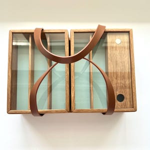 May include: A wooden cabinet with two sections, each with glass panels and interior dividers. A brown leather handle arches over the top and down the front. The cabinet has a light brown finish and a light green interior.
