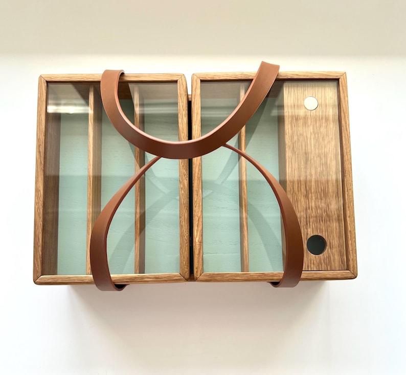 May include: A wooden cabinet with two sections, each with glass panels and interior dividers. A brown leather handle arches over the top and down the front. The cabinet has a light brown finish and a light green interior.