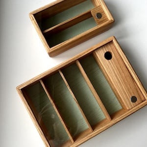 Puede incluir: Dos cajas de almacenamiento de madera con tapas transparentes. La caja más grande tiene cuatro compartimentos, mientras que la más pequeña tiene dos. Ambas cajas están hechas de madera de color claro y tienen un interior verde claro. Las cajas tienen un cierre magnético.