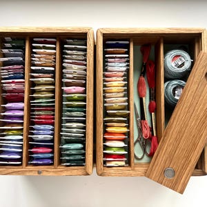 May include: A wooden embroidery floss organizer with multiple compartments filled with colorful thread spools. The open box reveals spools of thread, scissors, and embroidery floss. The lid is partially open, showing a circular handle.