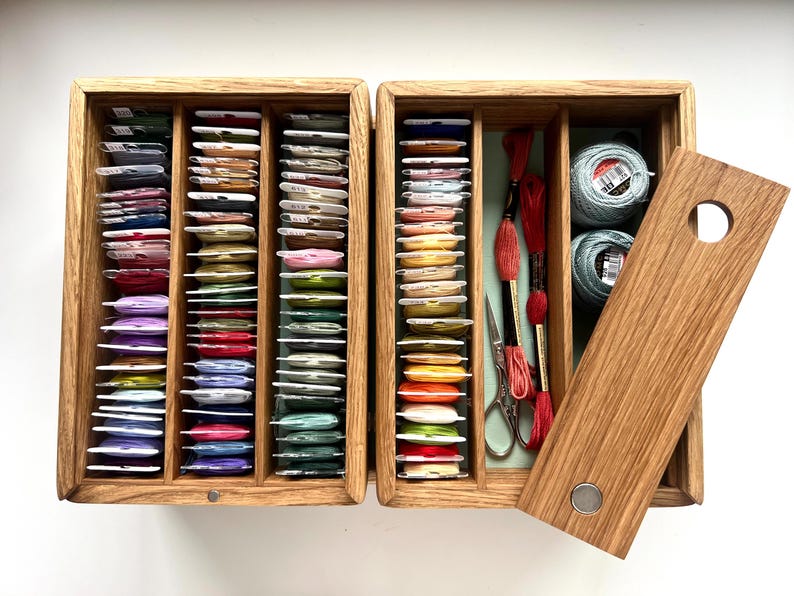 May include: A wooden embroidery floss organizer with multiple compartments filled with colorful thread spools. The open box reveals spools of thread, scissors, and embroidery floss. The lid is partially open, showing a circular handle.