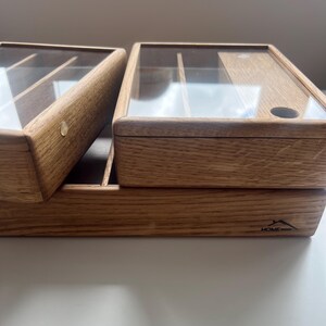 May include: Wooden storage box with a clear glass lid. The box is made of light brown wood and has a rectangular shape. The lid is hinged and opens to reveal compartments inside. The Home Wood logo is visible on the front.