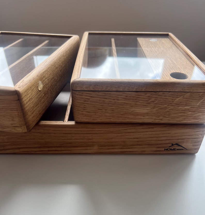 May include: Wooden storage box with a clear glass lid. The box is made of light brown wood and has a rectangular shape. The lid is hinged and opens to reveal compartments inside. The Home Wood logo is visible on the front.