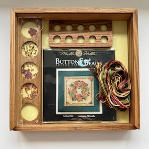 Puede incluir: Un kit de punto de cruz enmarcado en madera con el texto "Button & Beads Autumn Ser" y "Autumn Wreath". El kit incluye un patrón de punto de cruz, hilos coloridos y cuentas. El marco tiene un fondo amarillo.