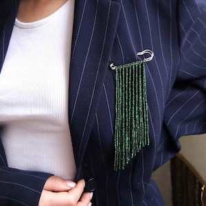 May include: A silver-toned safety pin brooch featuring multiple long, dangling strands of small, vibrant green beads. The brooch is attached to the lapel of a dark navy blue pinstriped blazer worn over a plain white ribbed tank top.