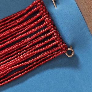 May include: A red beaded fringe accessory featuring multiple vertical strands of small, uniform red beads attached to a gold-toned metal safety pin at the top and a small gold-toned metal ring at the bottom corner, creating a linear pattern.
