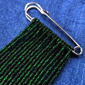 May include: A silver-toned metal safety pin featuring multiple vertical strands of small, dark green glass beads attached to the pin's bar. The beads are uniform in size and create a fringe-like appearance hanging from the metallic fastener.