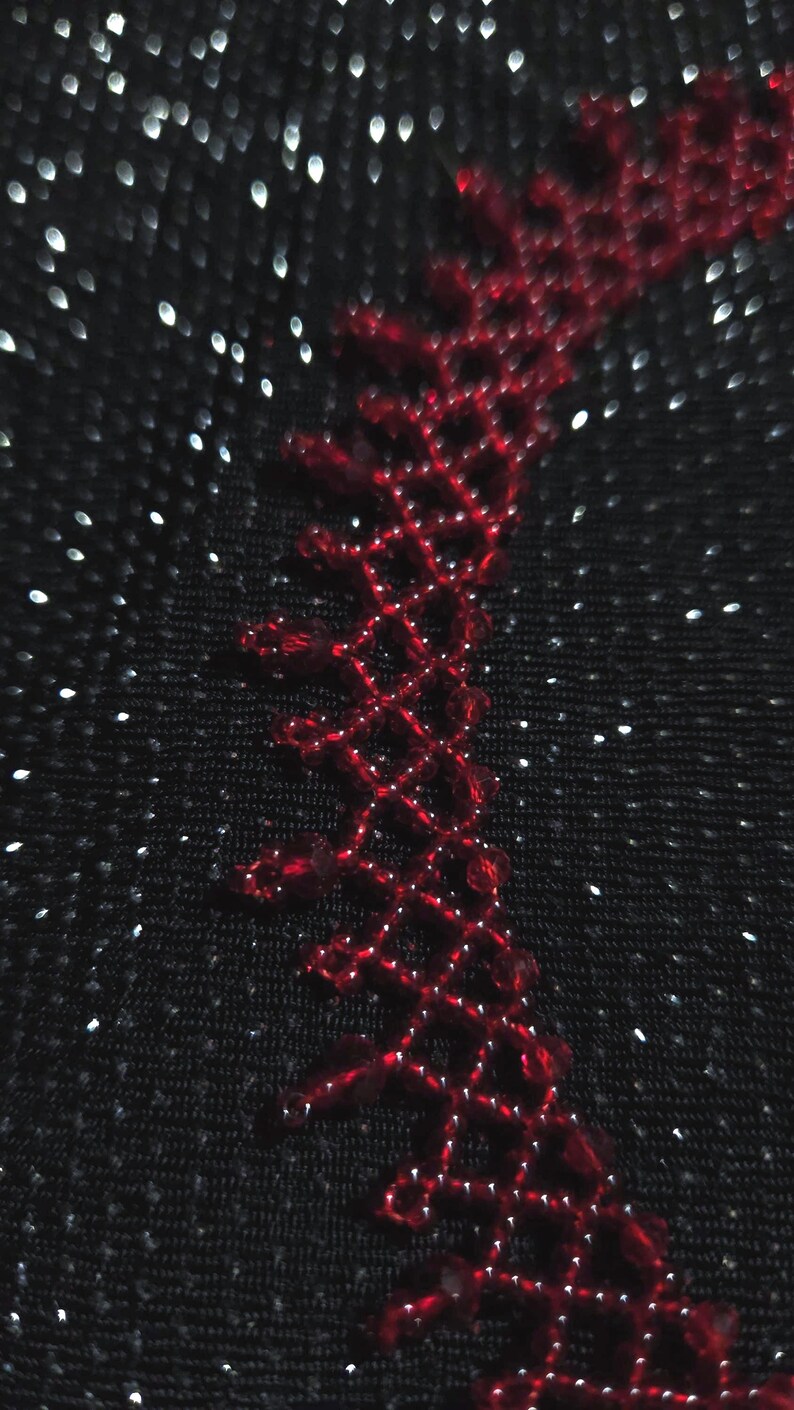 May include: Close-up of a black fabric with a red, open-weave design along the edge. The design is made of small, connected diamond shapes. The background has a subtle sparkle effect, with tiny white dots scattered across the surface.