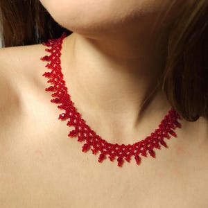 May include: A red beaded necklace with a delicate, intricate design. The necklace is made of small, round beads and sits close to the neck. The image shows the necklace on a person.