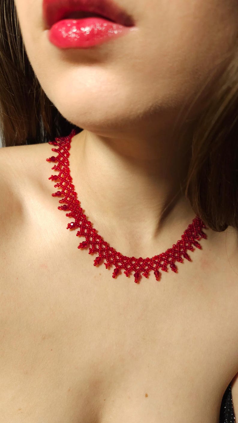 May include: A red beaded necklace with a delicate, intricate design. The necklace is made of small, round beads and sits close to the neck. The image shows the necklace on a person.