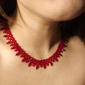May include: A red beaded necklace with a delicate, openwork design. The necklace is made of small, round beads and sits around the neck. The beads are a vibrant red color, creating a striking contrast.