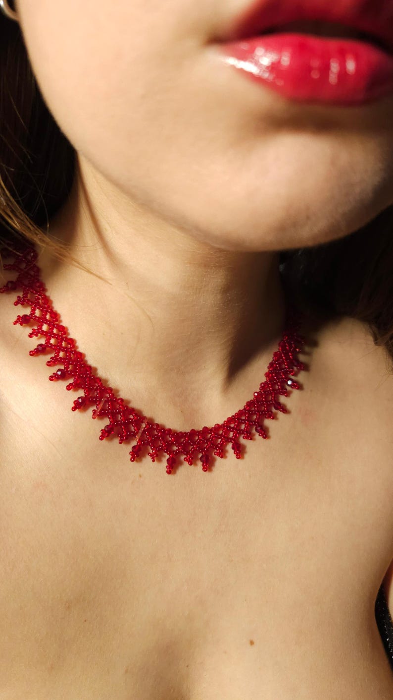 May include: A red beaded necklace with a delicate, openwork design. The necklace is made of small, round beads and sits around the neck. The beads are a vibrant red color, creating a striking contrast.