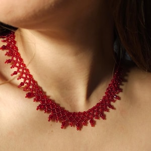 May include: A close-up of a red beaded necklace. The necklace is made of small, round beads and has a delicate, intricate design. The necklace is worn around the neck and sits just below the collarbone. The beads are a vibrant, solid red color.