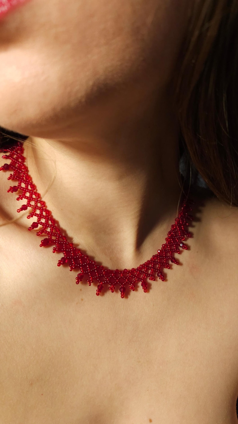 May include: A close-up of a red beaded necklace. The necklace is made of small, round beads and has a delicate, intricate design. The necklace is worn around the neck and sits just below the collarbone. The beads are a vibrant, solid red color.