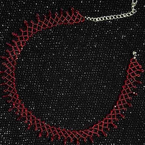 May include: A red beaded choker necklace with a delicate, openwork design. The necklace is made of small, red beads and features a silver-colored chain clasp. The background is a black, glittery fabric.