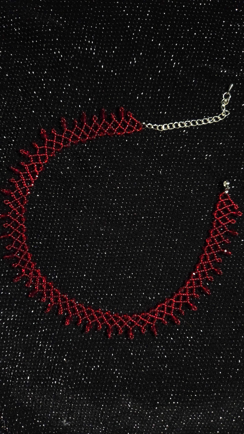 May include: A red beaded choker necklace with a delicate, openwork design. The necklace is made of small, red beads and features a silver-colored chain clasp. The background is a black, glittery fabric.