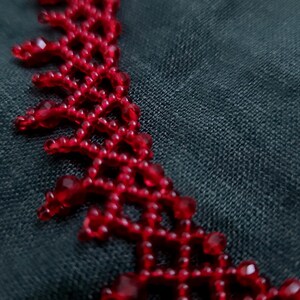 May include: A close-up of a red beaded necklace. The necklace features a delicate, openwork design with small, round beads and larger, faceted beads. The necklace is a vibrant red color and is set against a dark background.