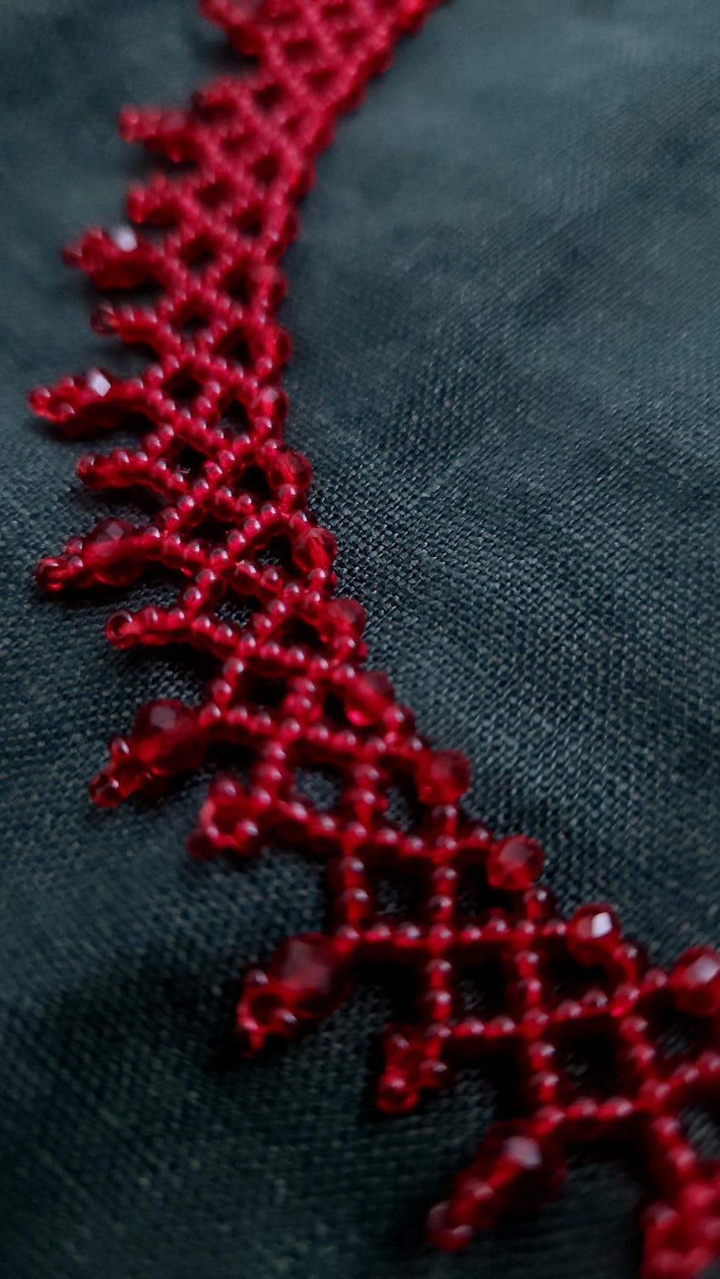 May include: A close-up of a red beaded necklace. The necklace features a delicate, openwork design with small, round beads and larger, faceted beads. The necklace is a vibrant red color and is set against a dark background.