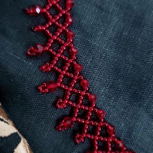 May include: Close-up of a red beaded necklace with a geometric design. The necklace is made of small, faceted beads and is set against a dark teal fabric background. The intricate beadwork creates a delicate, lacy effect.